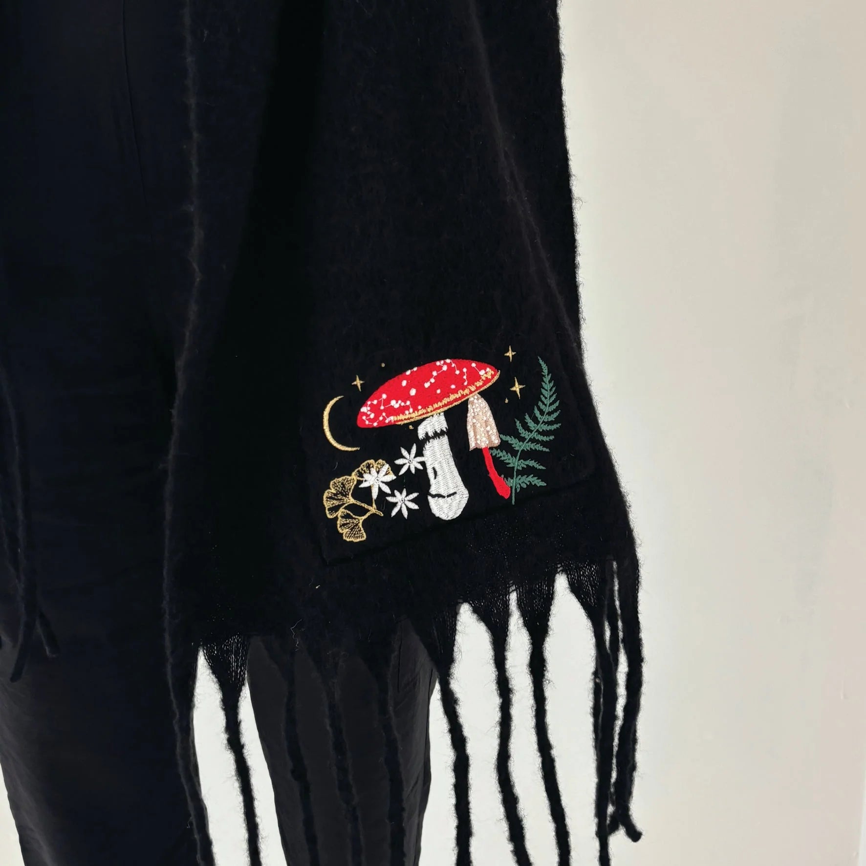 Close-up of bottom edge of Forage Scarf showing toadstool, fern, and flower embroidery against soft black knit fabric.