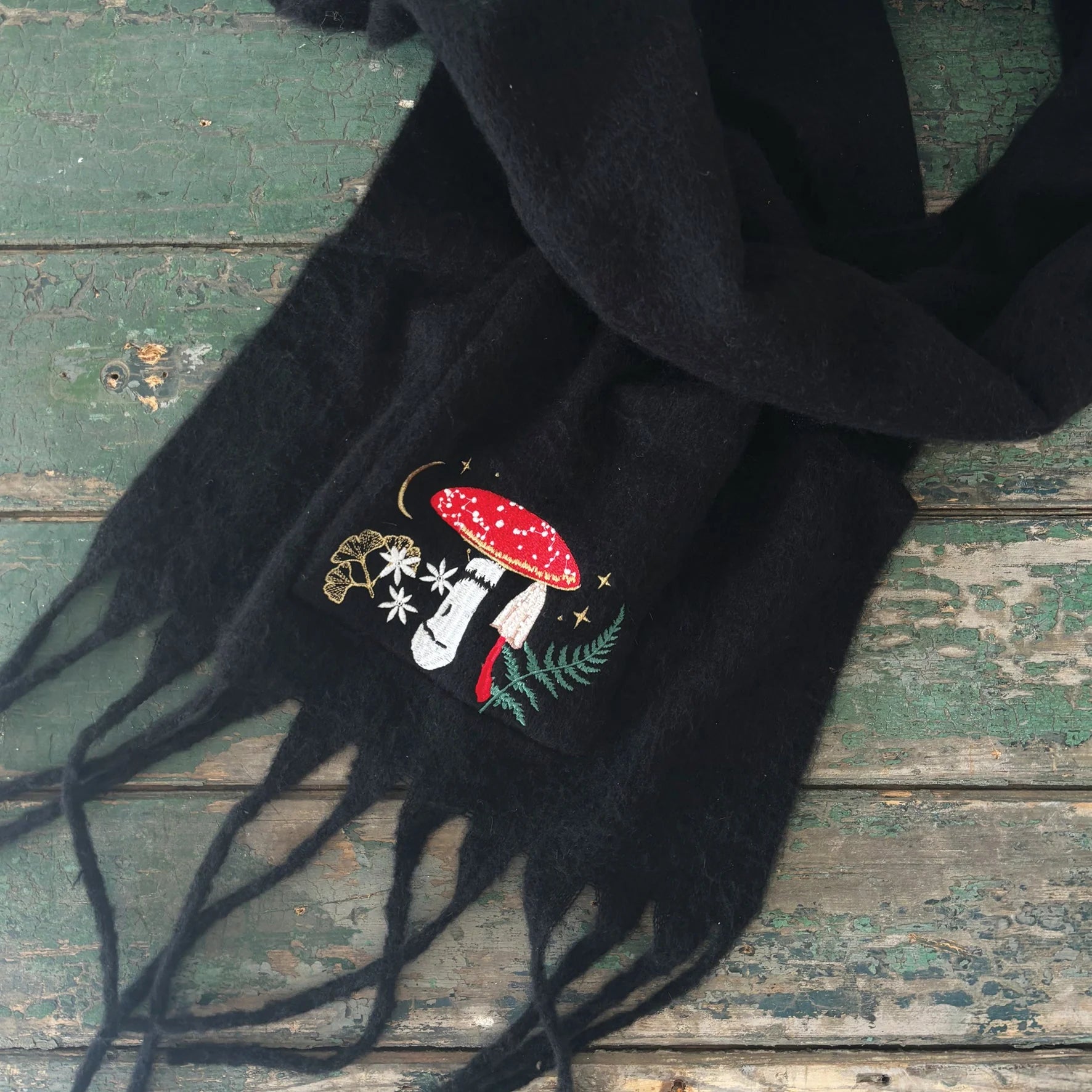 Flat lay of black Forage Scarf with fringe and detailed embroidered forest floor scene including a red toadstool, white flowers, fern, and gold stars.