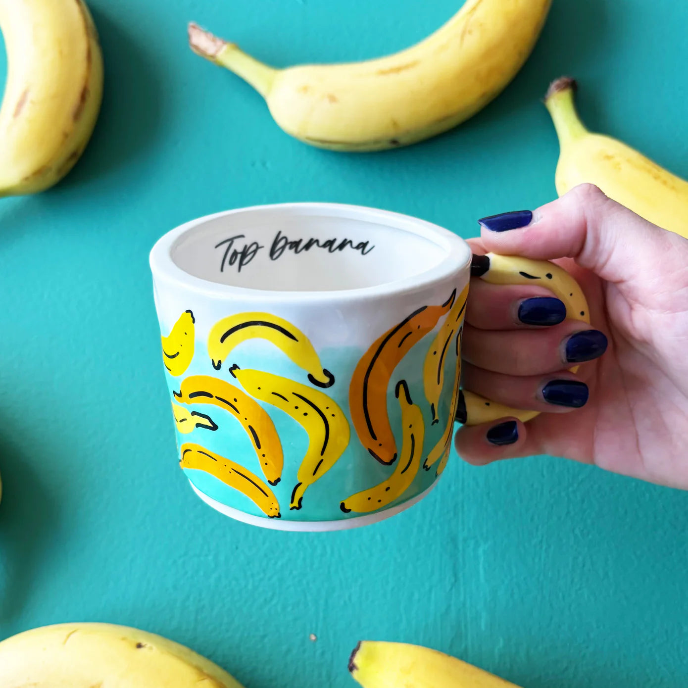 Front view of ceramic banana mug with curved 3D banana handle and colourful banana print on a teal background, “Top Banana” text visible inside.