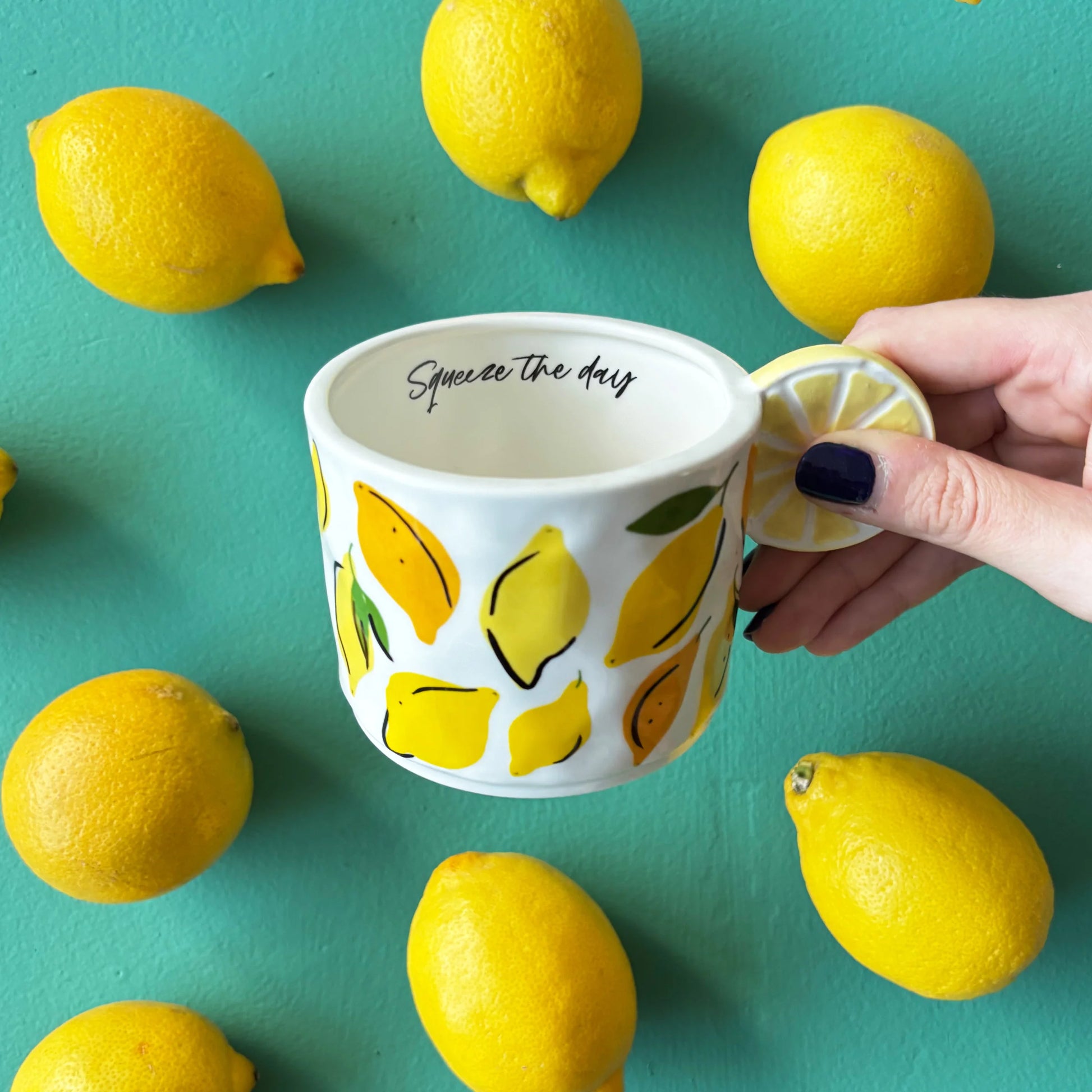 Hand holding ceramic lemon mug with 3D lemon slice handle, surrounded by real lemons on a turquoise background, showing “Squeeze the day” message inside.