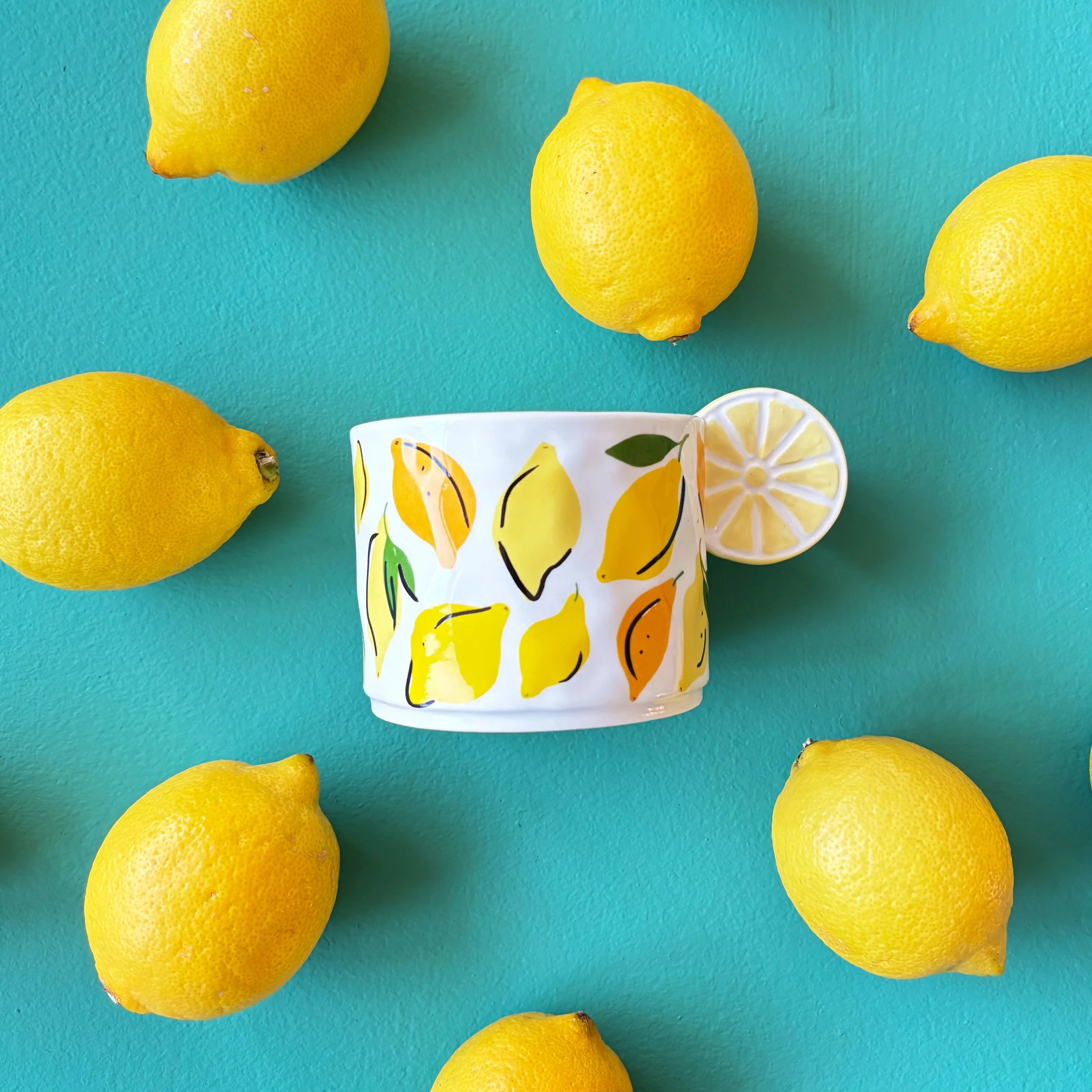 Top-down flat lay of fruity lemon mug with 3D slice handle, placed among fresh lemons on bright turquoise surface.