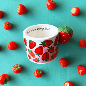 Flat lay of strawberry mug with illustrated pink design and strawberry-shaped handle, placed among scattered fresh strawberries.