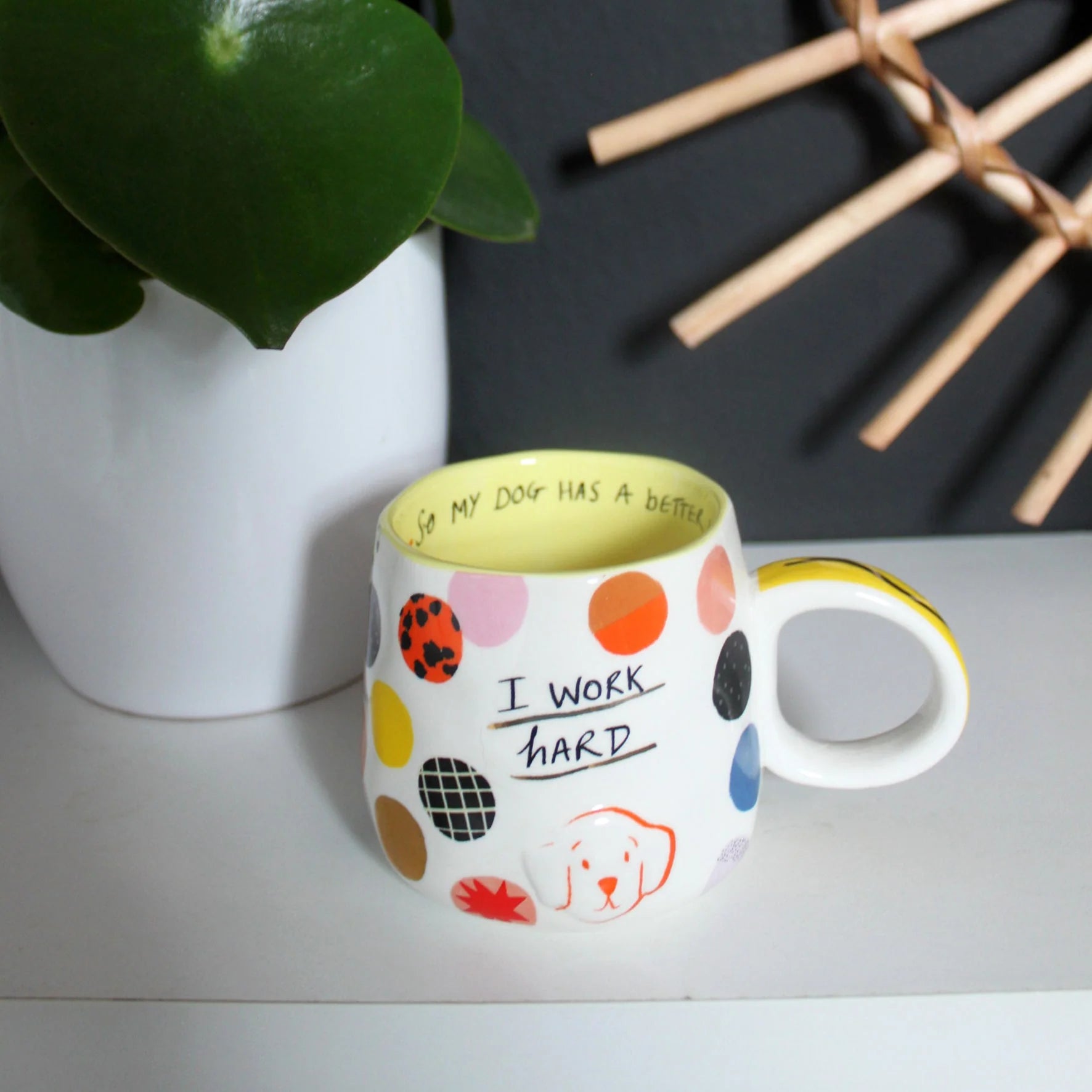 Quirky polka dot mug with yellow interior glaze and “I work hard” slogan, styled on a modern shelf next to a plant.