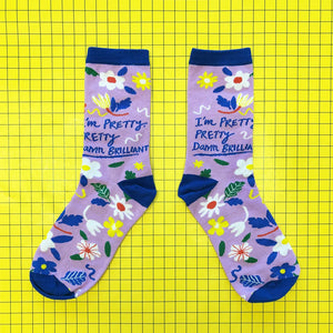 Flat lay of lilac bamboo socks with floral print and bold slogan “I’m pretty… pretty damn brilliant” on a yellow grid background.