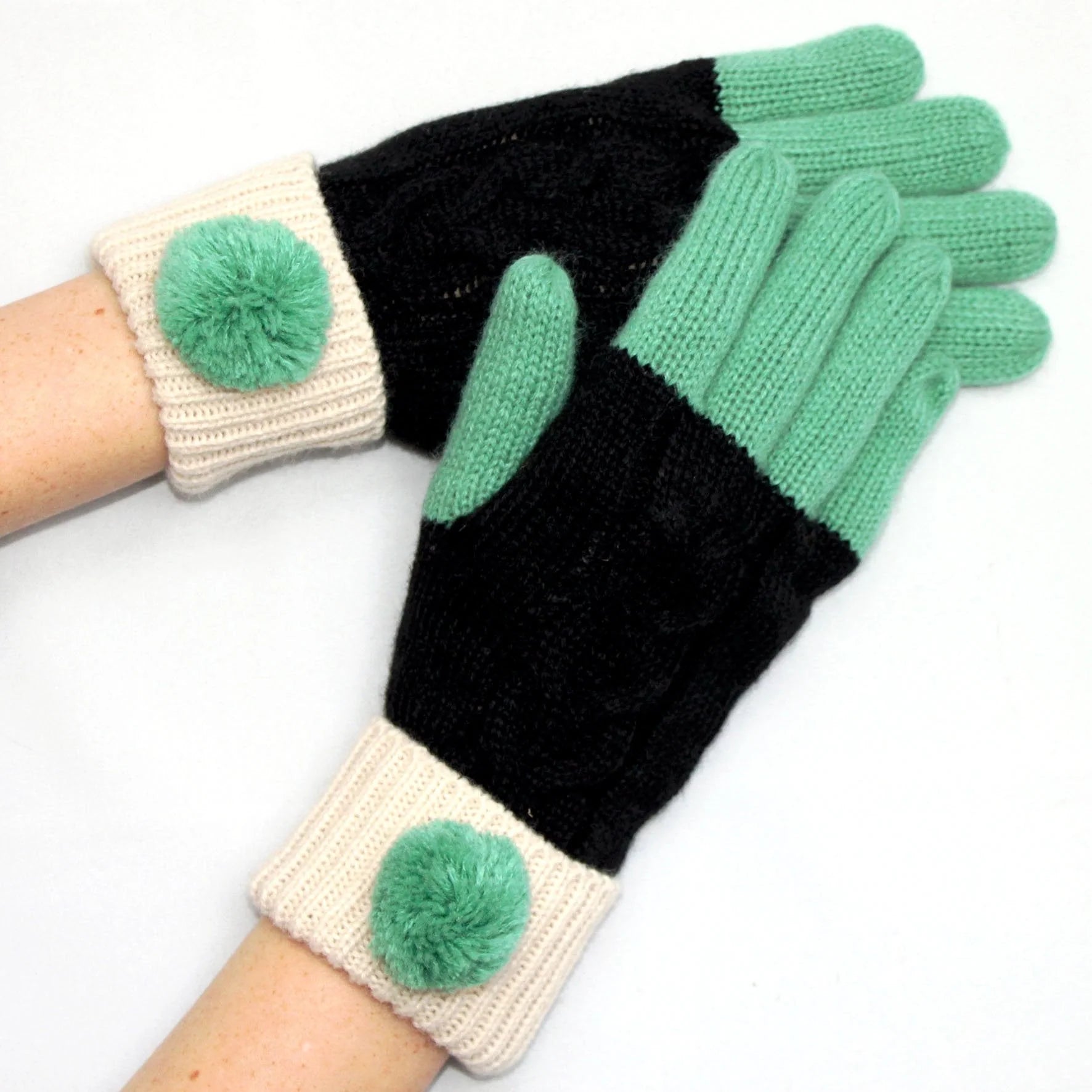 Hands wearing Loop Black Gloves showing colour-block knit design with mint fingers, black body, and cream cuffs with pom-poms.