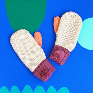 Pair of Loop Cream Mittens with orange thumbs and berry pom-pom cuffs displayed on a bold blue background with abstract shapes.