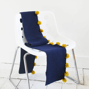 Full product view: Full length of navy scarf with mustard pom poms shown across white chair and floor.