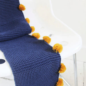 Detail view: Navy cable knit scarf with contrasting pom poms draped over white chair.
