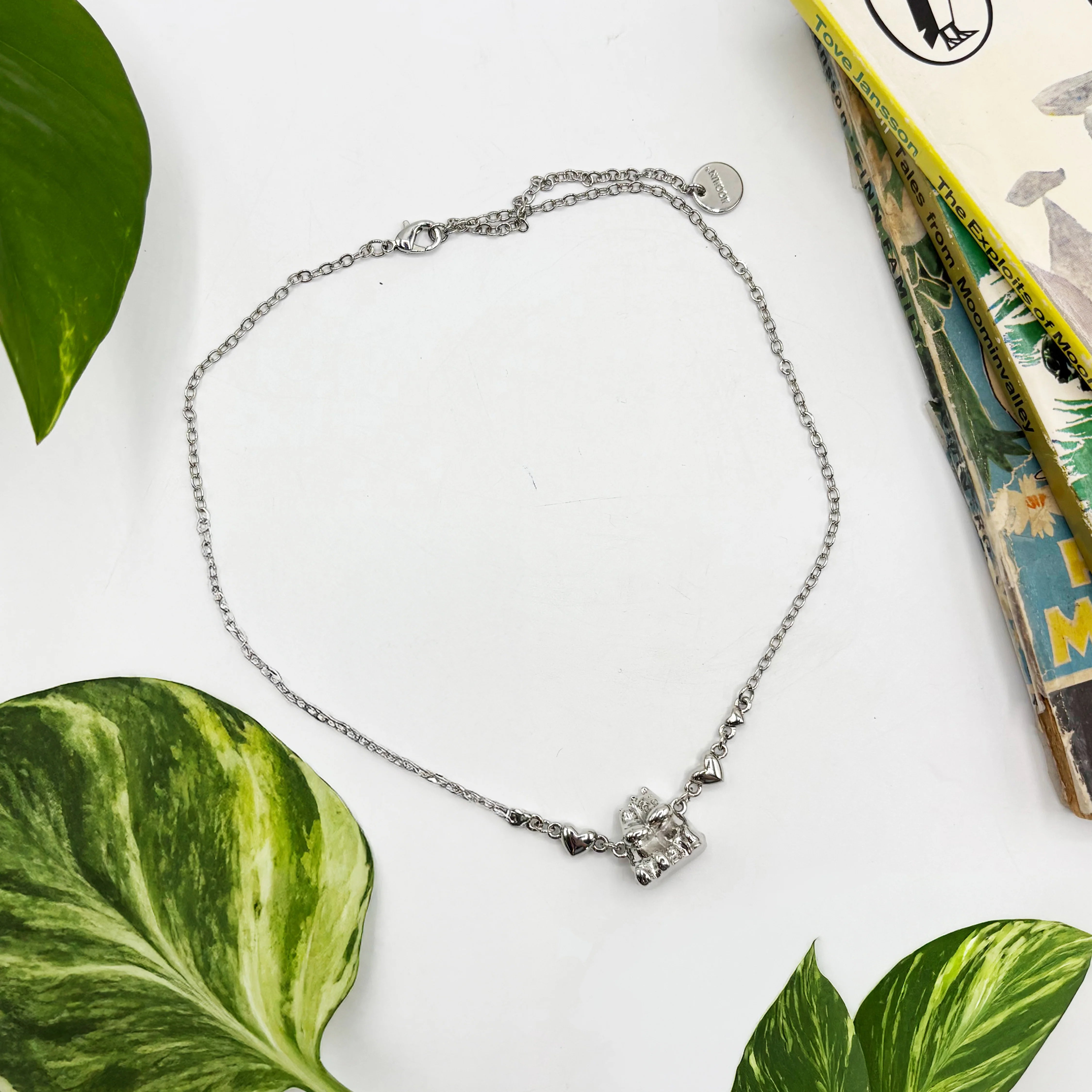 Silver Moomin love necklace laid flat with heart details and Moomintroll pendant, styled with plants and a book.
