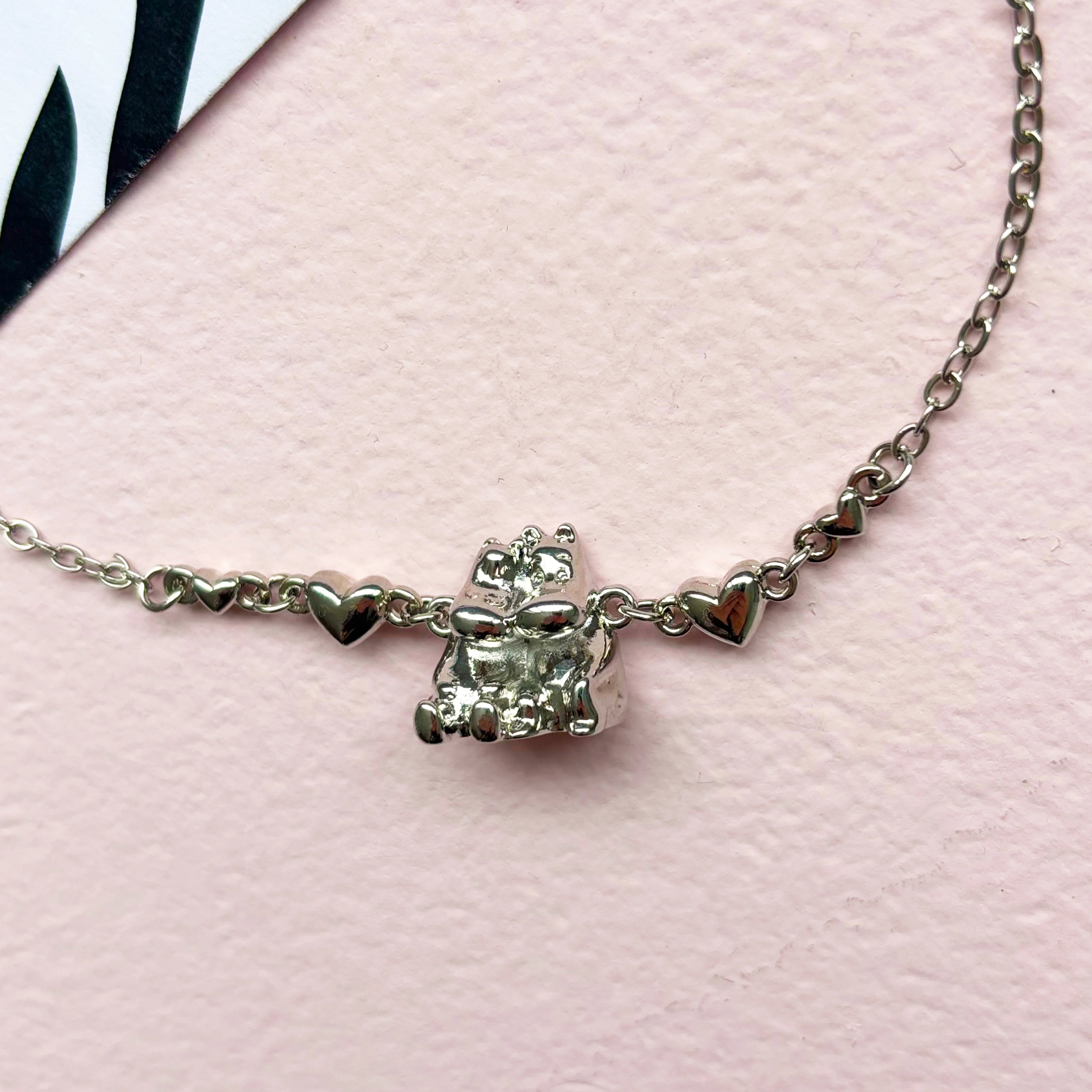 Close-up of the silver Moomin and Snorkmaiden hug pendant with heart chain links on pink background.