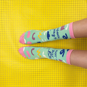 Person wearing mismatched slogan socks with bright pink toes and multicoloured pattern against yellow grid floor.