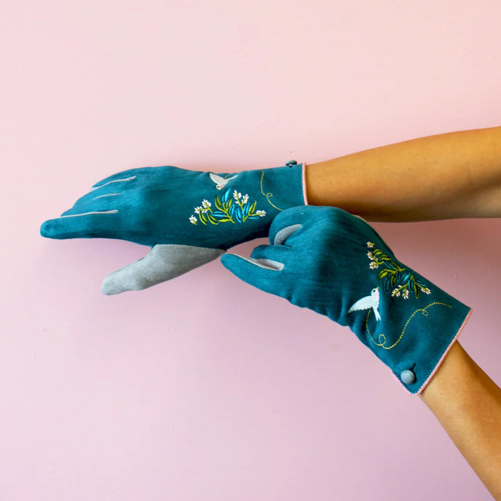 Model wearing teal embroidered bird gloves with grey fingertips and pink trim against a pale pink background.