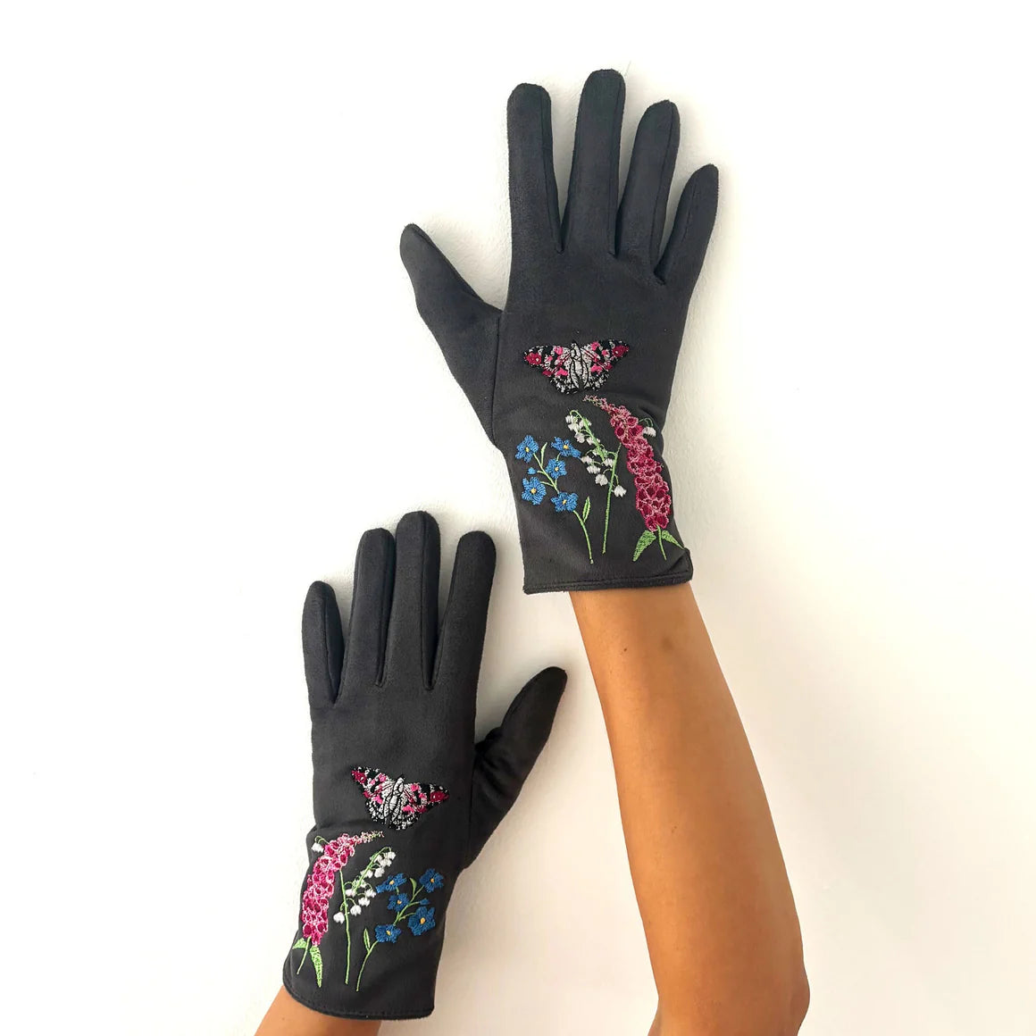 Top-down view of a person modelling grey embroidered gloves with colourful butterfly and wildflower design, displayed with one hand raised and the other relaxed.