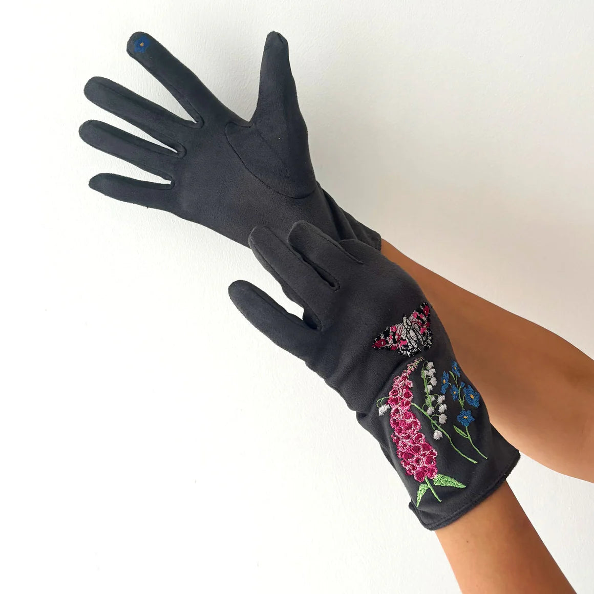 Angled view showing the top and inner palm of dark grey Secret Garden Butterfly Gloves, revealing floral embroidery and blue touchscreen detail on the thumb.