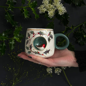 Side view of robin mug cradled in hand, highlighting holly berry design and gold foil accents.