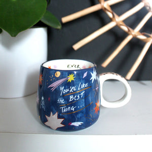 Ceramic mug with cosmic design and hand-lettered quote on shelf with plant decor.
