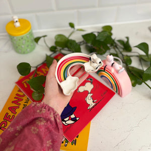 A hand holding the Snoopy Rainbow Trinket Dish, with Peanuts-themed books and greenery in the background.