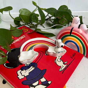 The Snoopy Rainbow Trinket Dish placed on a red Peanuts book, next to a matching rainbow-themed Snoopy decoration and green plants.