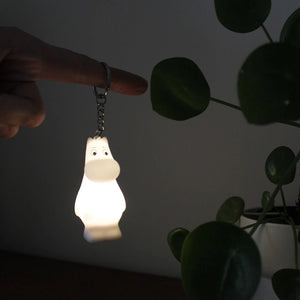 A hand holding a glowing Moomin light-up keyring in a dark setting, with a plant in the background.