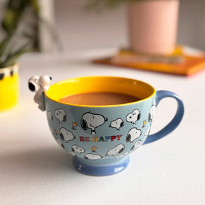 A vibrant blue and yellow Snoopy-themed mug filled with a hot beverage, featuring an all-over Snoopy and Woodstock print with “Be Happy” text and a 3D Snoopy figurine hanging off the side.