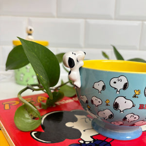 Close-up of the Snoopy ‘Be Happy’ Cup, showcasing the 3D Snoopy figurine attached to the side and surrounded by green plants, with a Peanuts comic book in the background.