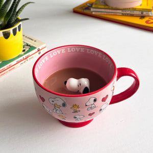  pink and red Peanuts Love Mug filled with tea, featuring Snoopy and Woodstock illustrations with heart details and a 3D Snoopy figurine inside.