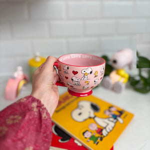 A person holding the Peanuts Love Mug with an all-over Snoopy and Woodstock print, hearts, and “LOVE” text along the rim.