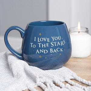 The I love you to the stars and back blue ceramic mug sitting on a rug and with a candle in the background.