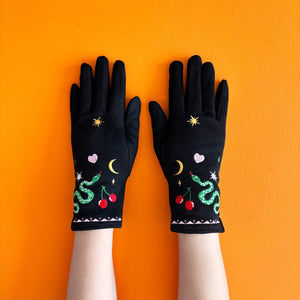 Black suedette embroidered gloves with snake, cherry, and heart designs, displayed flat on a bright orange background.