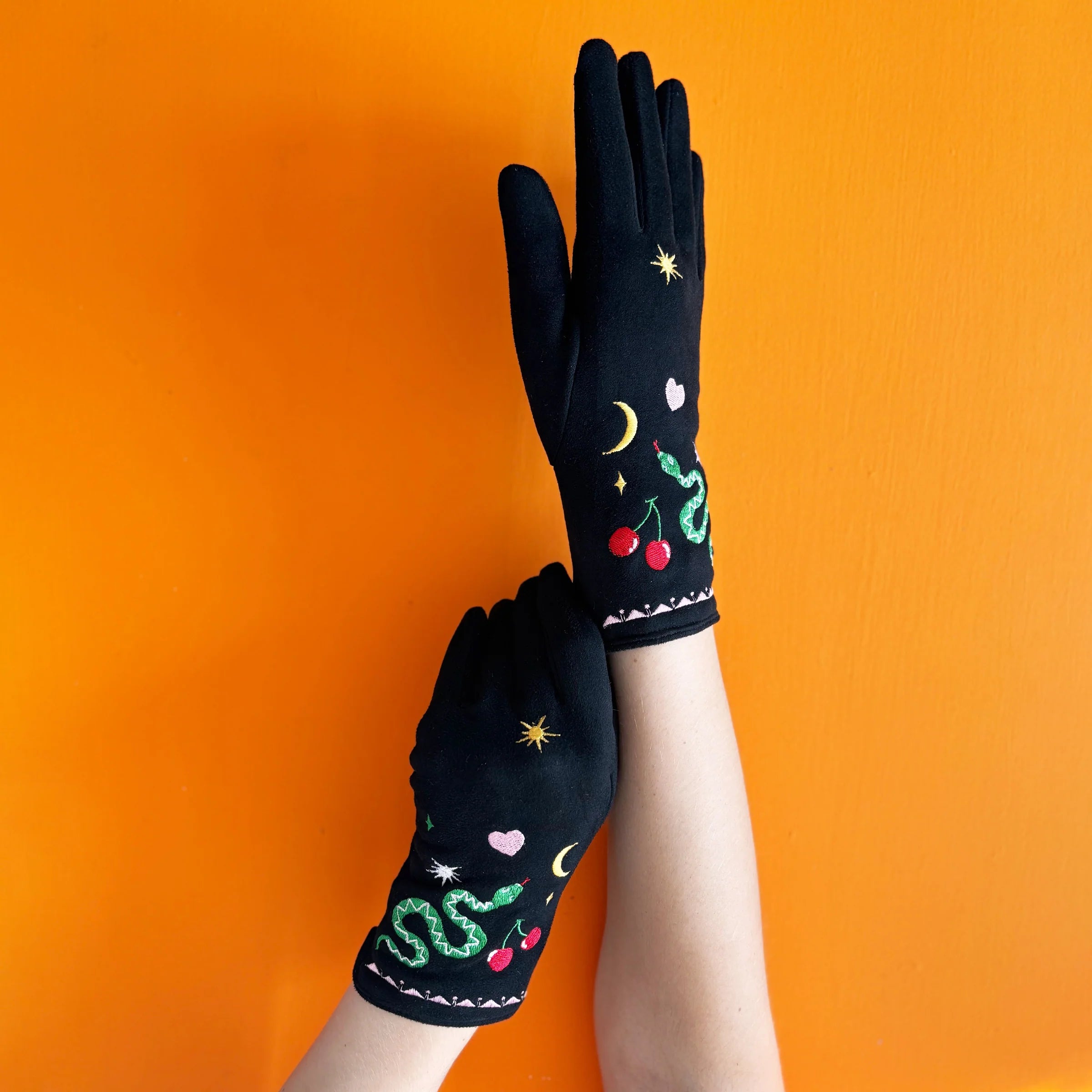 Hands wearing black embroidered snake gloves with cherries, moons, and hearts against an orange wall.