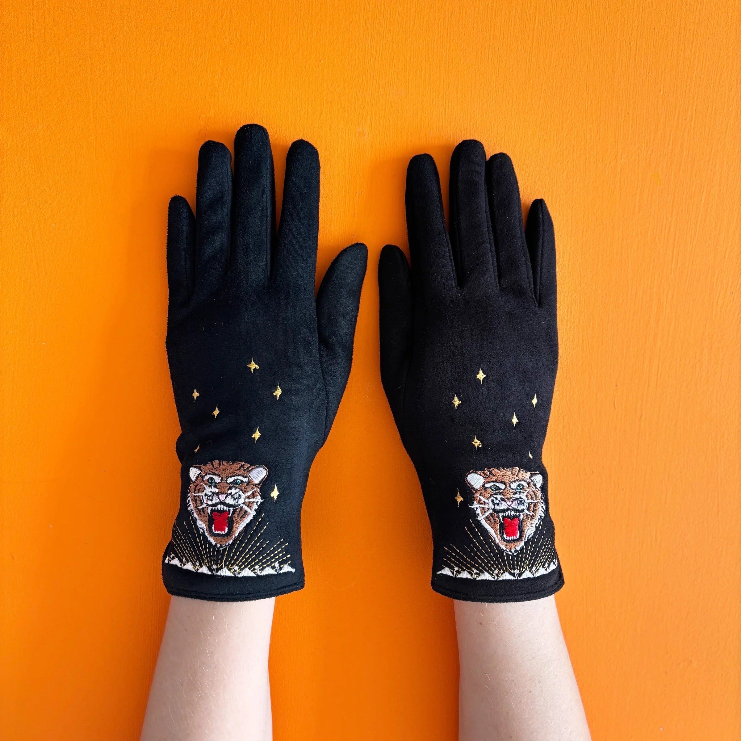 Black suedette embroidered tiger gloves with gold stars, displayed flat on a bright orange background.