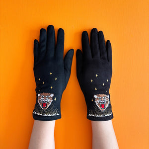 Black suedette embroidered tiger gloves with gold stars, displayed flat on a bright orange background.