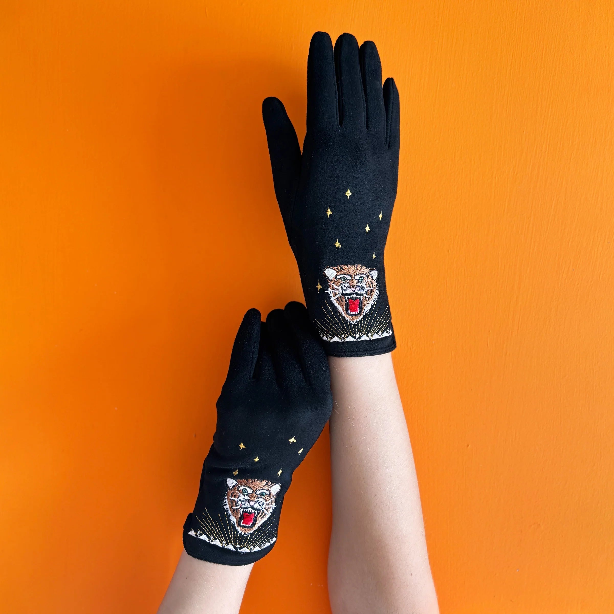 Hands wearing black embroidered tiger gloves with gold star details, shown against an orange wall.
