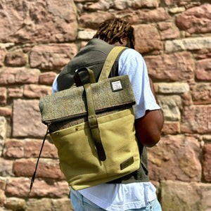 A man with a beard, wearing a blue sweater and jeans, is carrying a brown Islander Harris tweed canvas backpack.