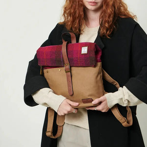 A woman with long, red hair, wearing a black coat and heels, is holding a red tartan Islander Harris tweed canvas backpack.