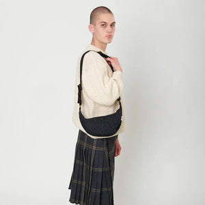 A bald woman wearing a kilt and white jumper is carrying a navy over-check Harris Tweed Luna bag over her shoulder.