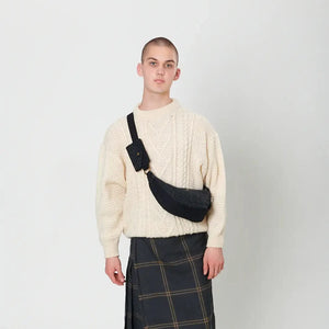 A bald woman wearing a kilt and white jumper is holding a navy over-check Harris Tweed Luna bag in front of her.