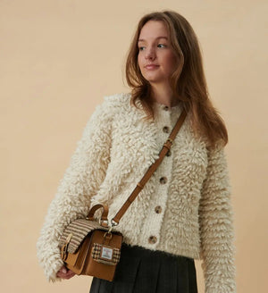 A woman wearing a beige Harris Tweed Melrose Leather Crossbody Bag over her shoulder. She is standing outdoors and smiling. The bag is made from genuine leather and features a traditional Harris Tweed pattern.