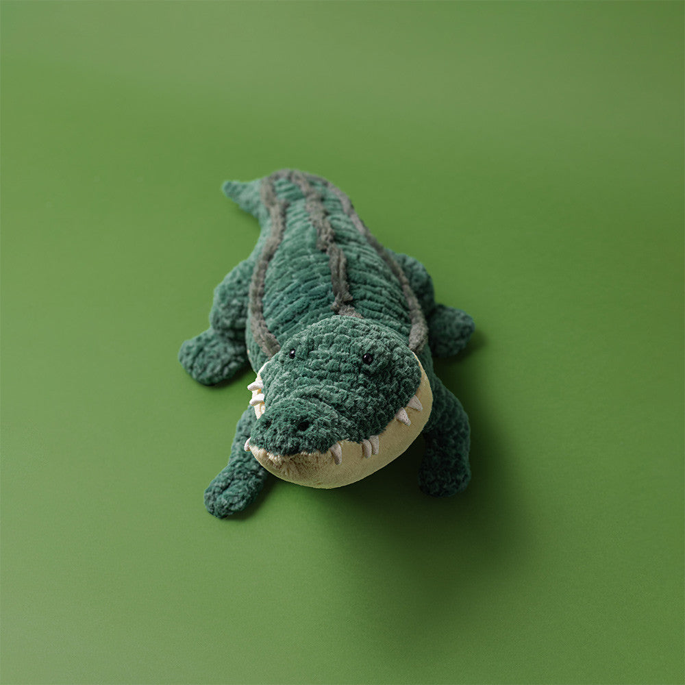 Jellycat Allexi Alligator plush toy photographed from above on a green background, showing its soft textured green body, corded ridged back, and friendly toothy grin.