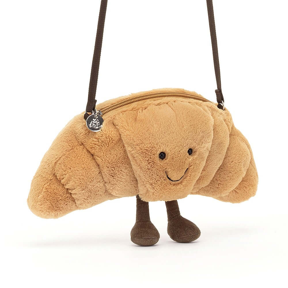 Unexpected Adventure: The Jellycat Amuseable Croissant Bag hanging from a tree branch in a park, suggesting its versatility for outdoor explorations and adventures.