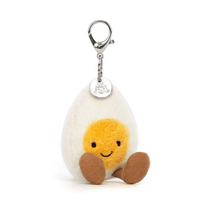 Jellycat Amuseable Happy Boiled Egg Bag Charm in a seated position, has a silver clip, the egg has soft cream fur and a sunny yellow yolk, brown corded boots and Jellycat Tag.