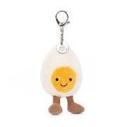 Jellycat Amuseable Happy Boiled Egg Bag Charm, has a silver clip, the egg has soft cream fur and a sunny yellow yolk, brown corded boots and Jellycat Tag.