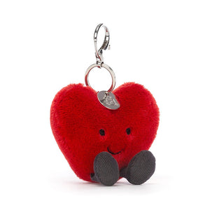 Jellycat Amuseable Heart Bag Charm in a sitting position with plush red fur, brown corded boots. It has a silver claw clip and the silver Jellycat tag.