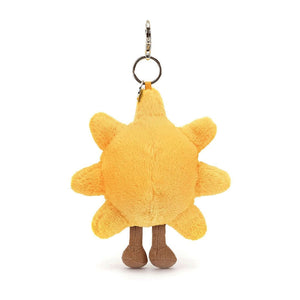 A Jellycat Amuseable Sun Bag Charm hanging from a bright blue backpack. The charm features a smiling sun with big black eyes, a golden plush body, and milk chocolate corduroy boots. It's attached to the backpack with a silver chain and claw clip.