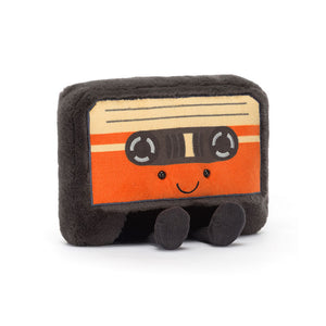 Angled View: Rewind to fun! The Jellycat Amuseables Cassette Tape leans in, showcasing its soft black fur, embroidered silver reels, and printed orange and butterscotch label. A blast from the past for little music lovers & cuddle enthusiasts!