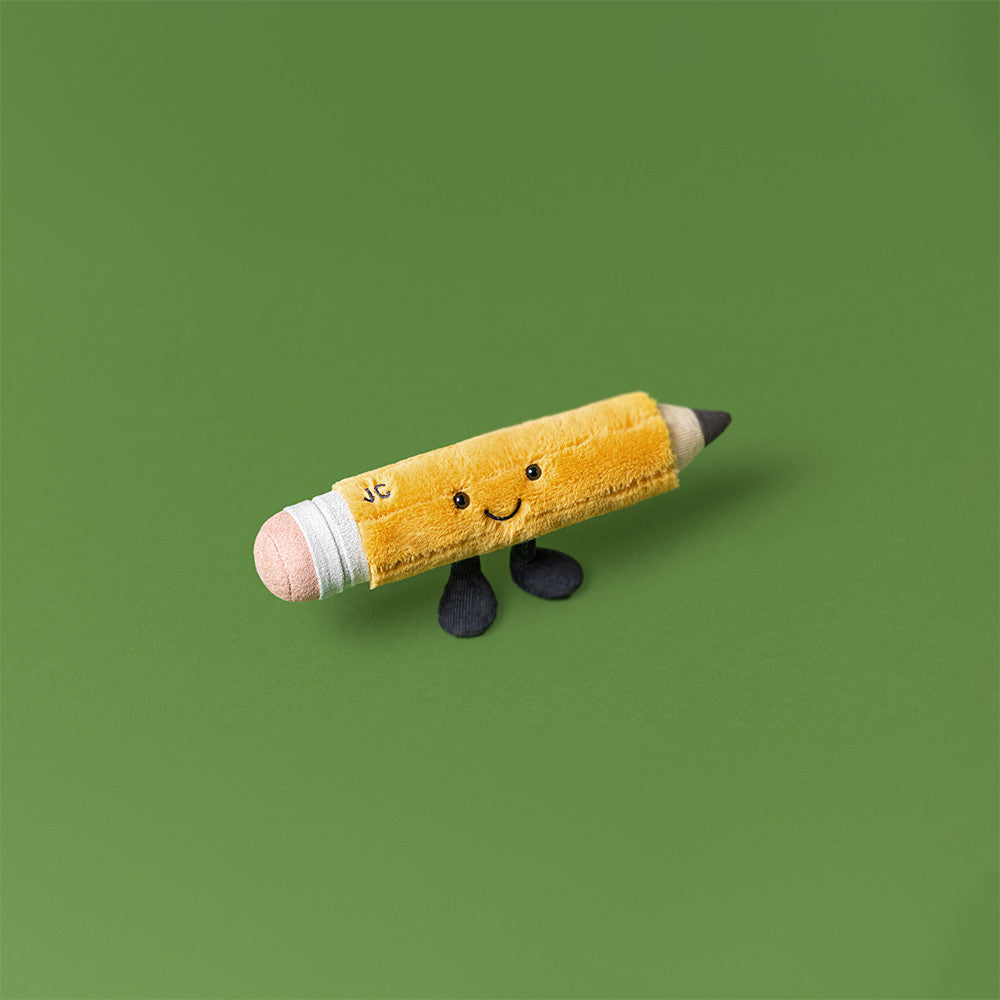 Playful Jellycat Amuseables Pencil plush toy posed upright on a green background, with stitched JC monogram, smiling embroidered face, and black corduroy legs