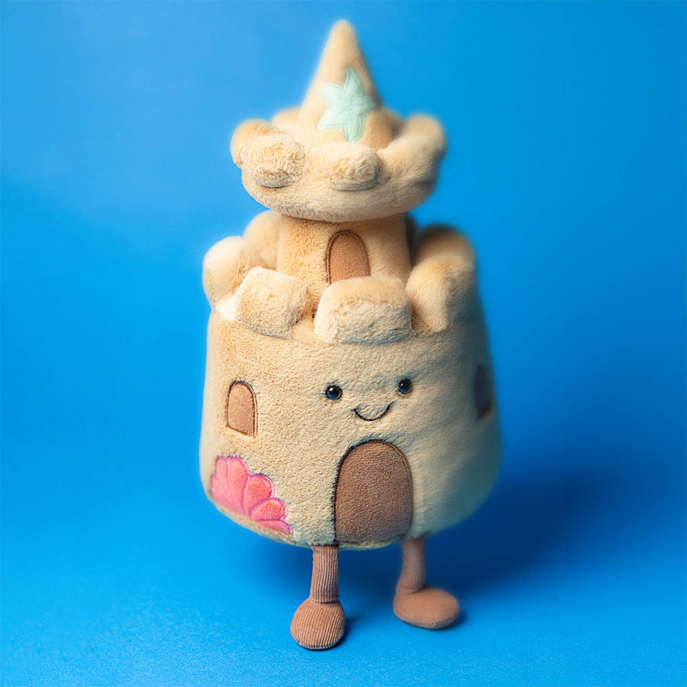 Jellycat Amuseables Sandcastle soft toy standing on a blue background with starfish detail and cheerful embroidered face.