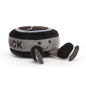 Angled View: Ready for playtime! The Jellycat Amuseables Sports Ice Hockey Puck leans in, showcasing its soft black fur, grey tummy band, denim-blue patches, and embroidered hockey sticks. Score playtime fun with this cuddly puck!