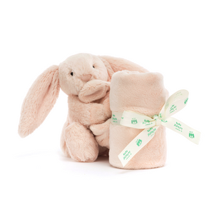 Jellycat Bashful Blush Bunny Soother. Soft blush pink bunny clutches the corner of a luxuriously soft, recycled plush blanket in calming blush pink, held together with Jellycat Ribbon.