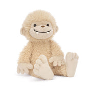 A buttercream Jellycat Bucky Bigfoot peeks sideways, showcasing his scruffy fur, oversized ears, big grin, and adorable bobbled toes.